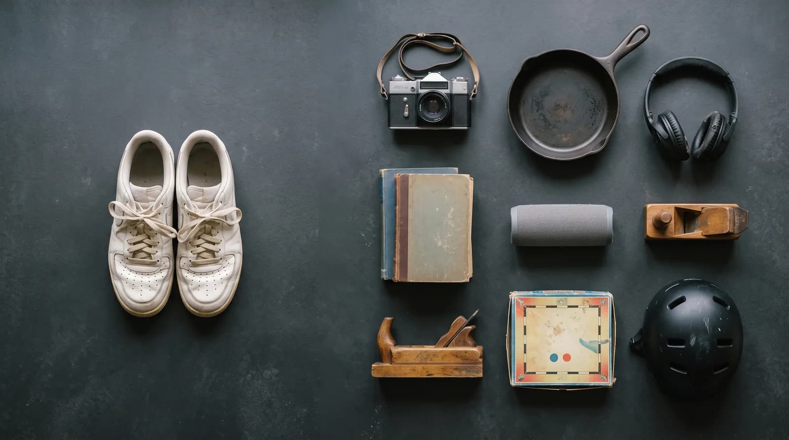 A pair of white trainers on the left of a dark slate surface, and nine other second-hand objects arranged opposite them: a film camera, a cast-iron pan, headphones, books, a speaker, two wooden hand planes, a boxed game and a skate helmet.
