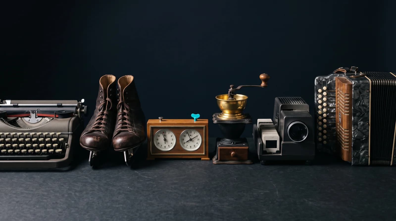 Six second-hand objects standing in a row on a dark surface: a portable typewriter, a pair of leather ice skates, a chess clock, a hand-cranked coffee grinder, a slide projector and a button accordion.