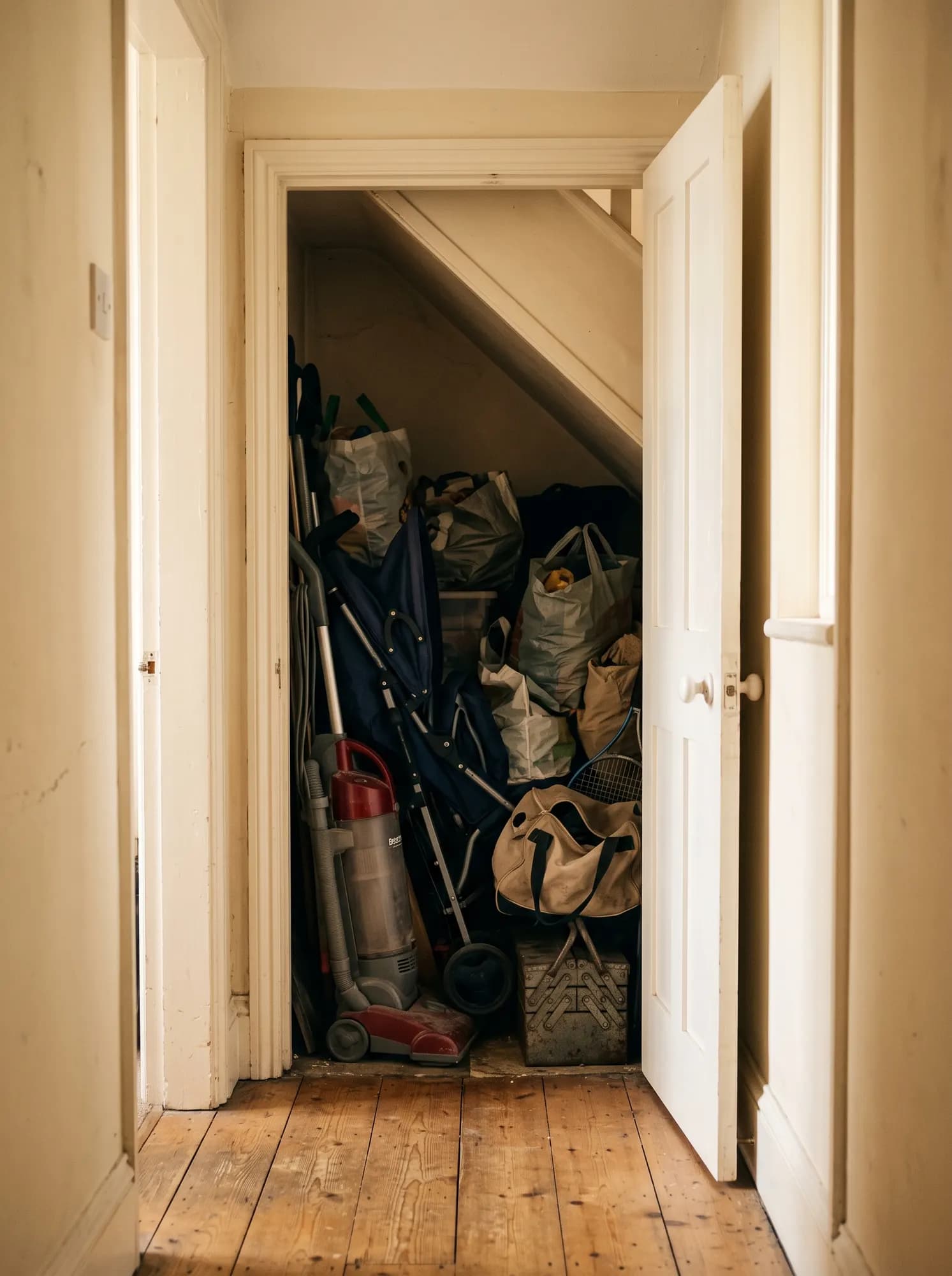 An under-stairs cupboard seen through an open door: an upright vacuum cleaner, a folded pushchair, a tennis racquet, a canvas holdall and carrier bags stacked to the sloped ceiling.