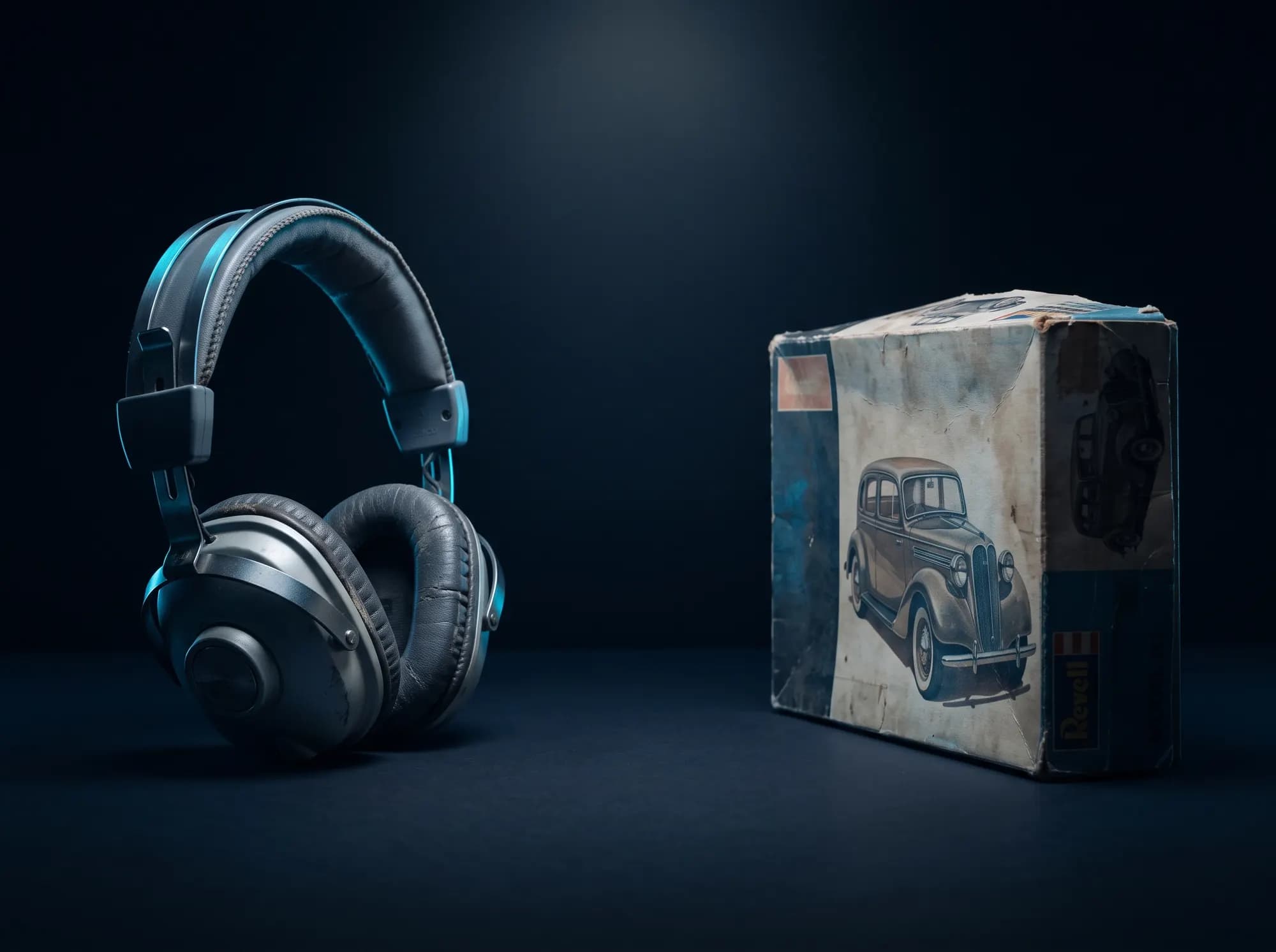 A pair of headphones and a boxed model kit facing each other across a dark surface, an empty gap between them.