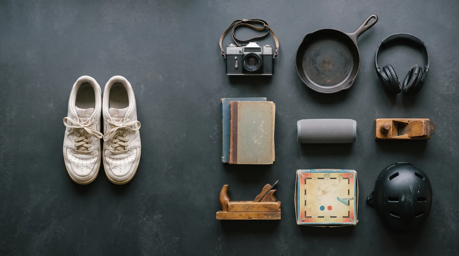 A pair of white trainers on the left of a dark slate surface, and nine other second-hand objects arranged opposite them: a film camera, a cast-iron pan, headphones, books, a speaker, two wooden hand planes, a boxed game and a skate helmet.